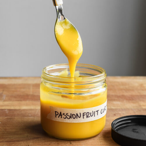 Passion fruit curd in a labeled jar with a spoon dipping.